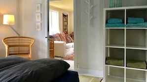 A bedroom with a bed, a wicker chair, a bedside table with a lamp, a wardrobe with shelves, and a door leading to another room.