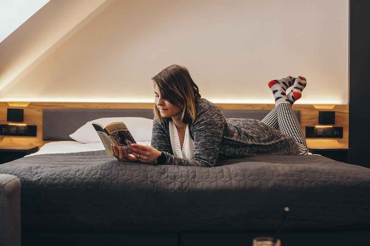 Eine Person liegt in einem Zimmer mit schrägwandiger Decke auf einem Bett und liest ein Buch.