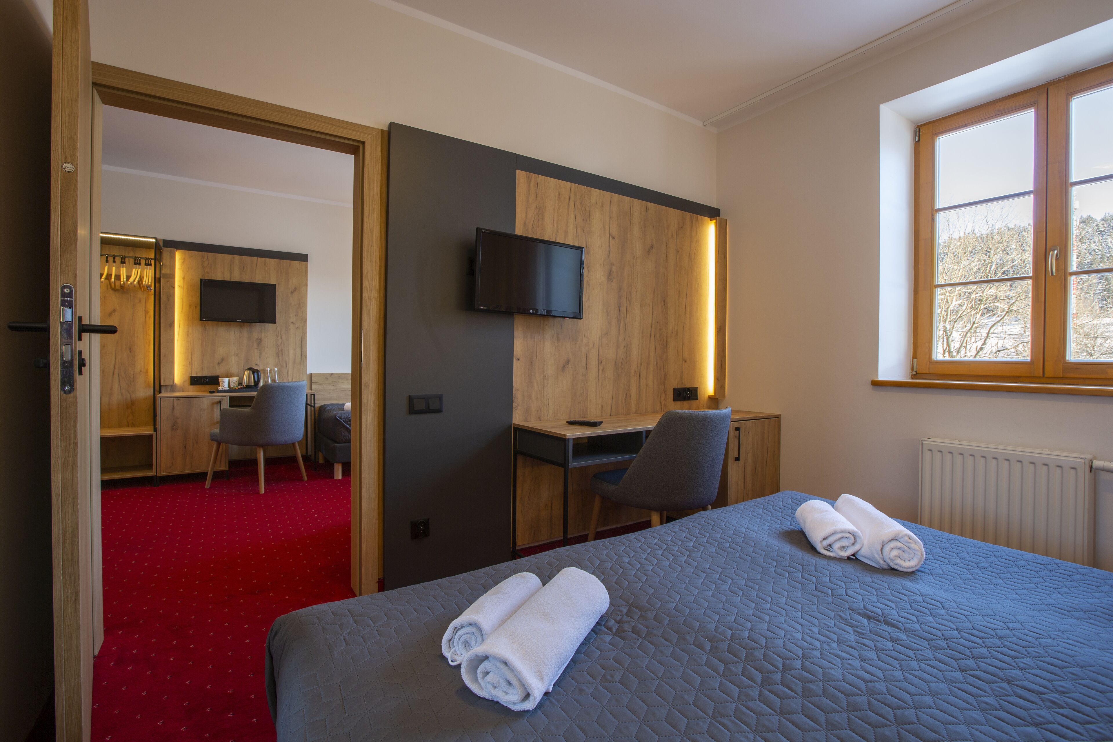 A hotel room with a bed, a desk, a chair, a TV mounted on the wall, and a window with a view of trees.