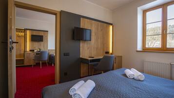 A hotel room with a bed, a desk, a chair, a TV mounted on the wall, and a window with a view of trees.