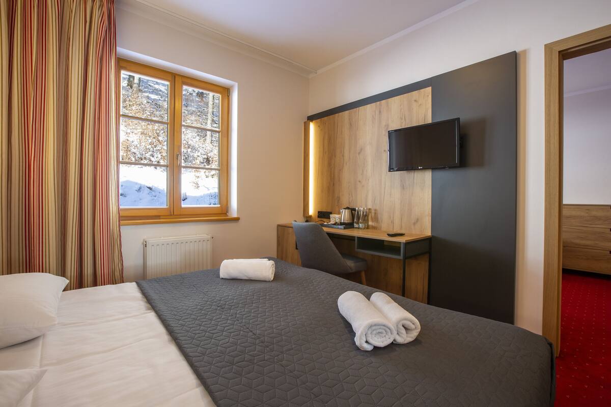 Ein Hotelzimmer mit Bett, Schreibtisch, Stuhl, Fernseher und einem Fenster mit Blick auf Bäume.