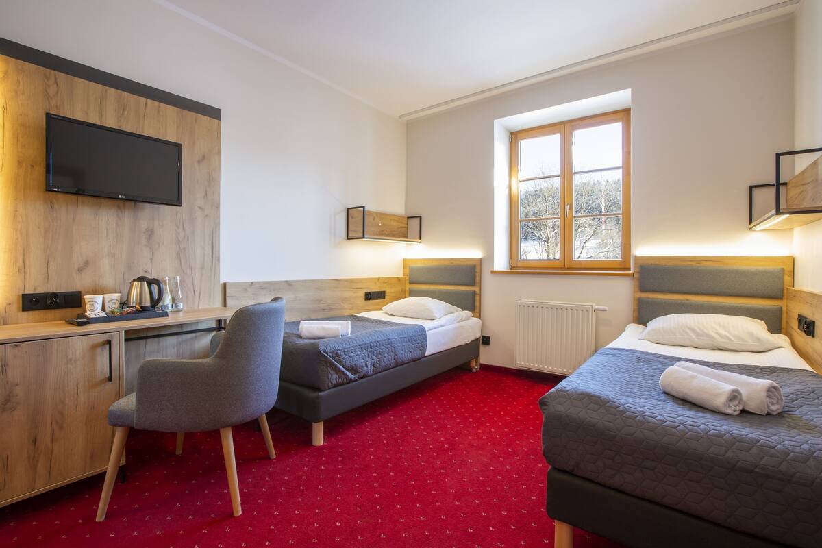 Ein Hotelzimmer mit zwei Betten, einem Schreibtisch, einem Stuhl, einem Fernseher und einem Fenster.