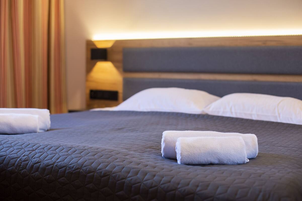 Ein ordentlich bezogenes Bett mit dunkler Decke, weißen Kissen, einer Nachttischlampe und einem gestreiften Vorhang.