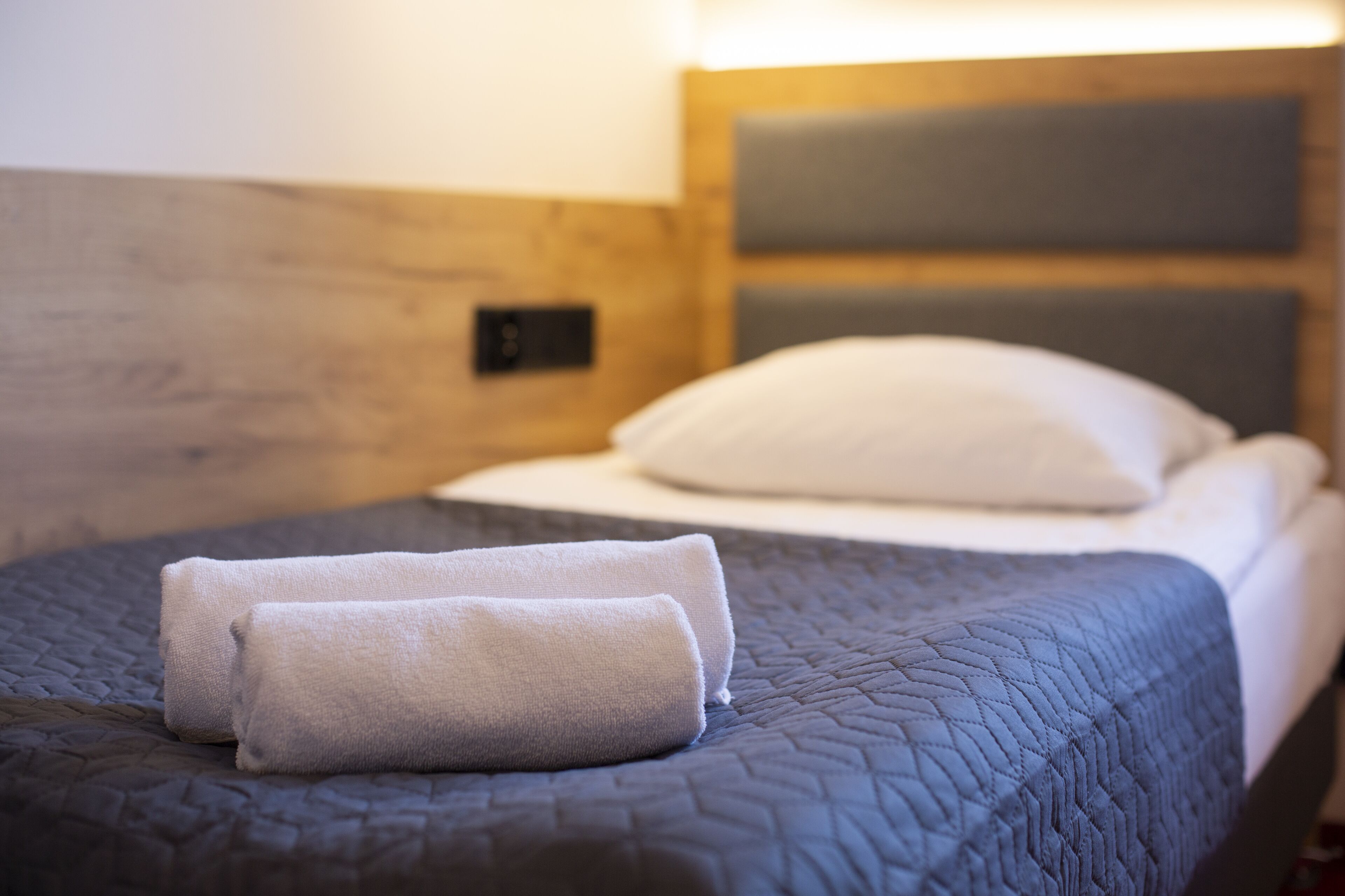 A neatly made bed with a dark quilt, two rolled towels, and a white pillow.