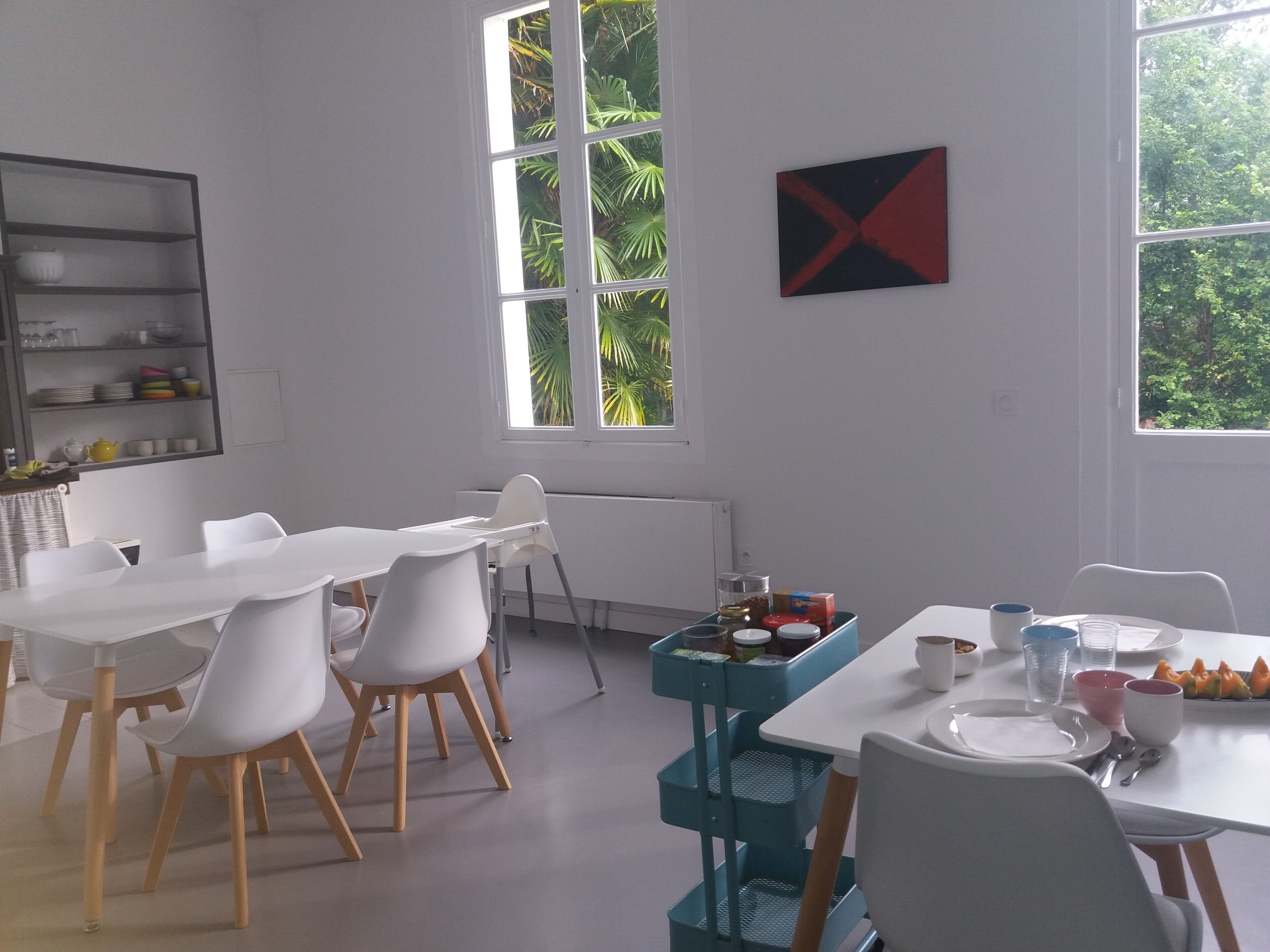 Un comedor con mesas y sillas blancas, un carrito azul con comida, y una ventana con vista a árboles.