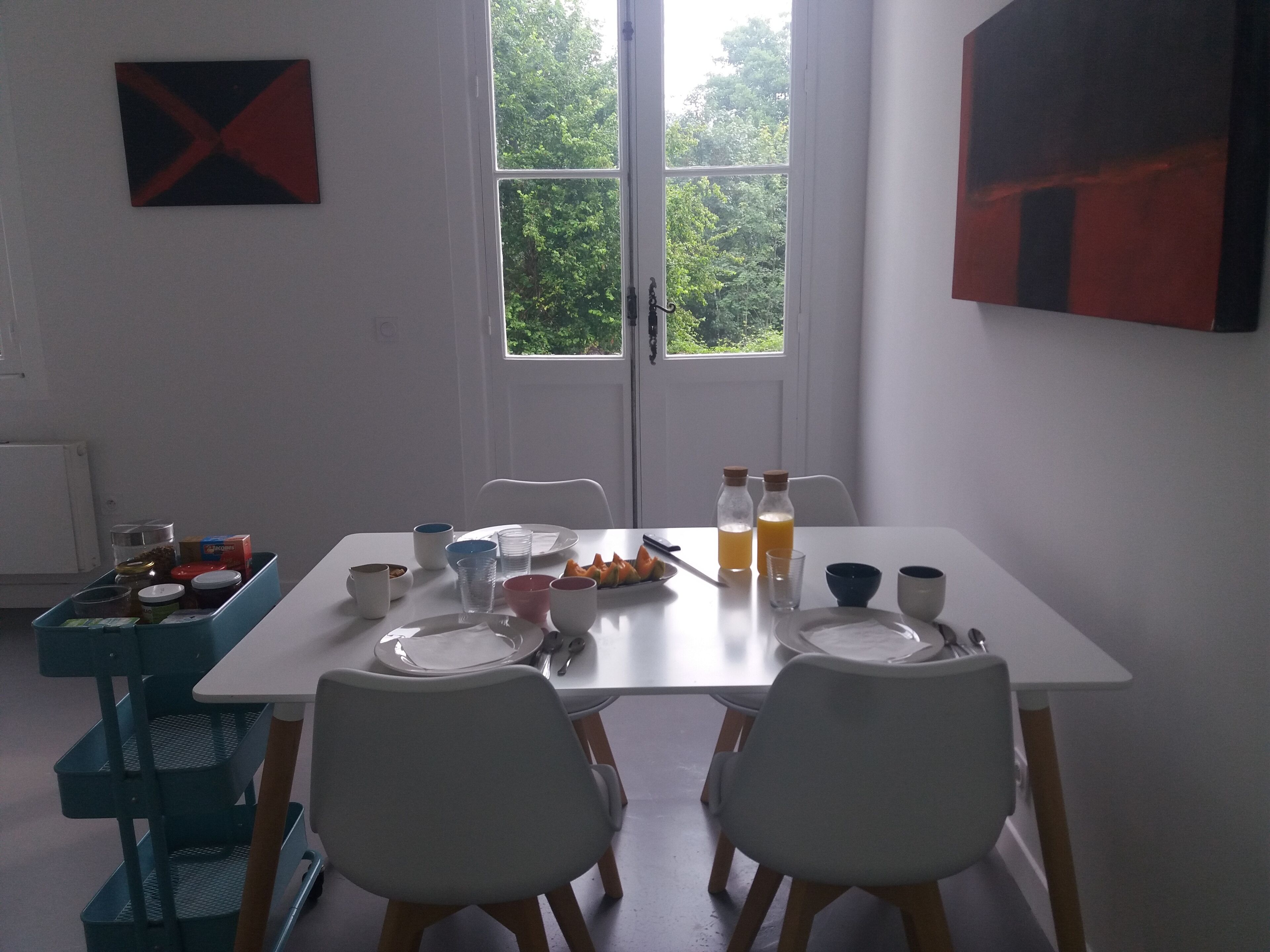 Un comedor con una mesa blanca dispuesta para dos personas, sillas, una ventana con vistas a zonas verdes y un mueble azul con contenedores.