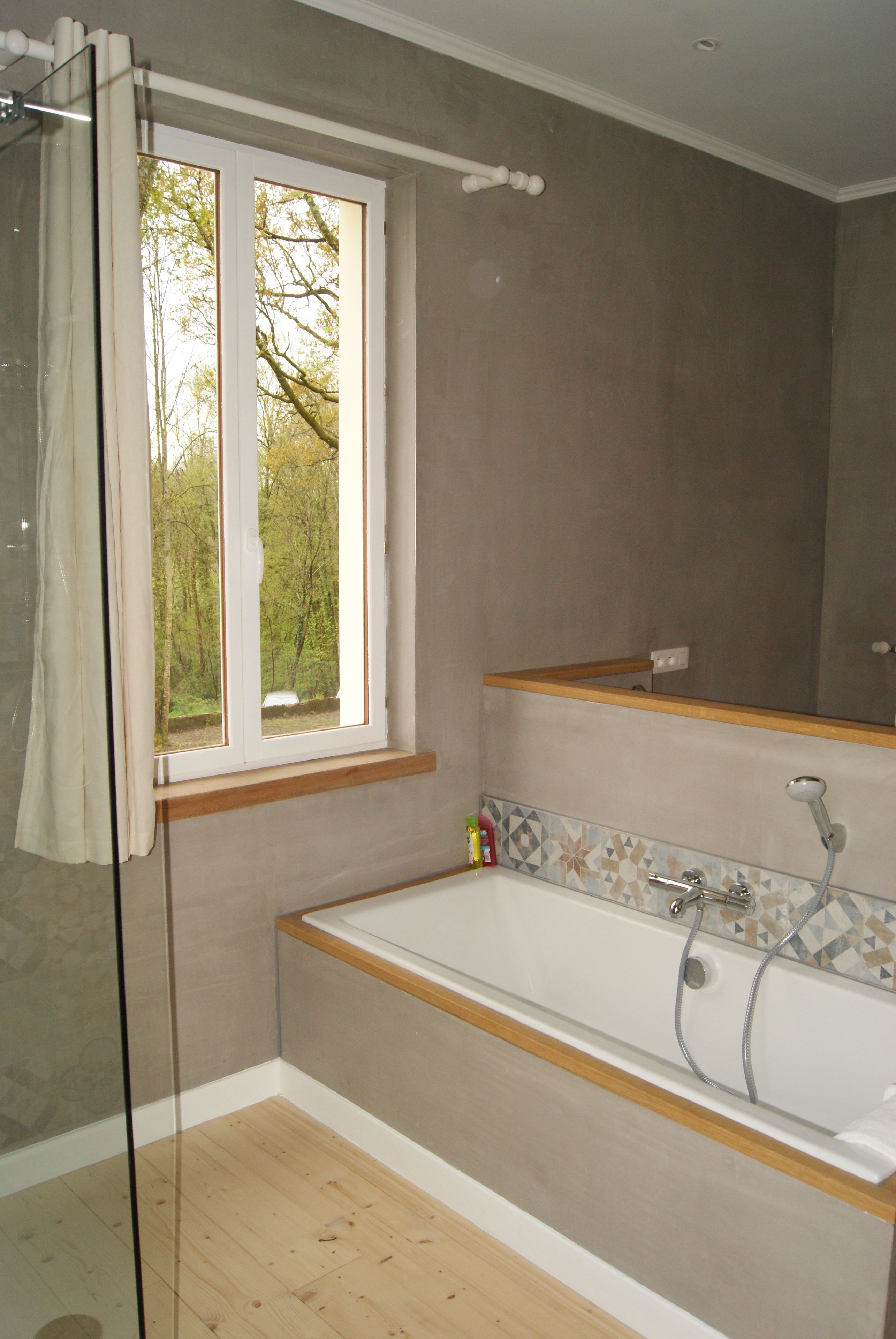 Un baño con bañera, ventana con vista a árboles y zona de ducha.