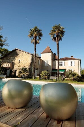 Zona junto a la piscina con dos grandes esculturas metálicas, un edificio de piedra con techo de tejas y palmeras.