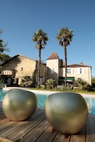 Zona junto a la piscina con dos grandes esculturas metálicas, un edificio de piedra con techo de tejas y palmeras.