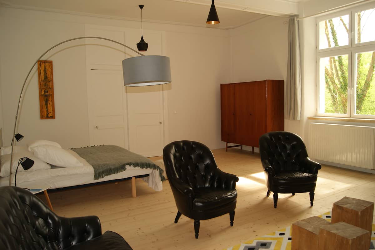 A bedroom with a bed, two black leather chairs, a wooden cabinet, and a large window with a view of trees.