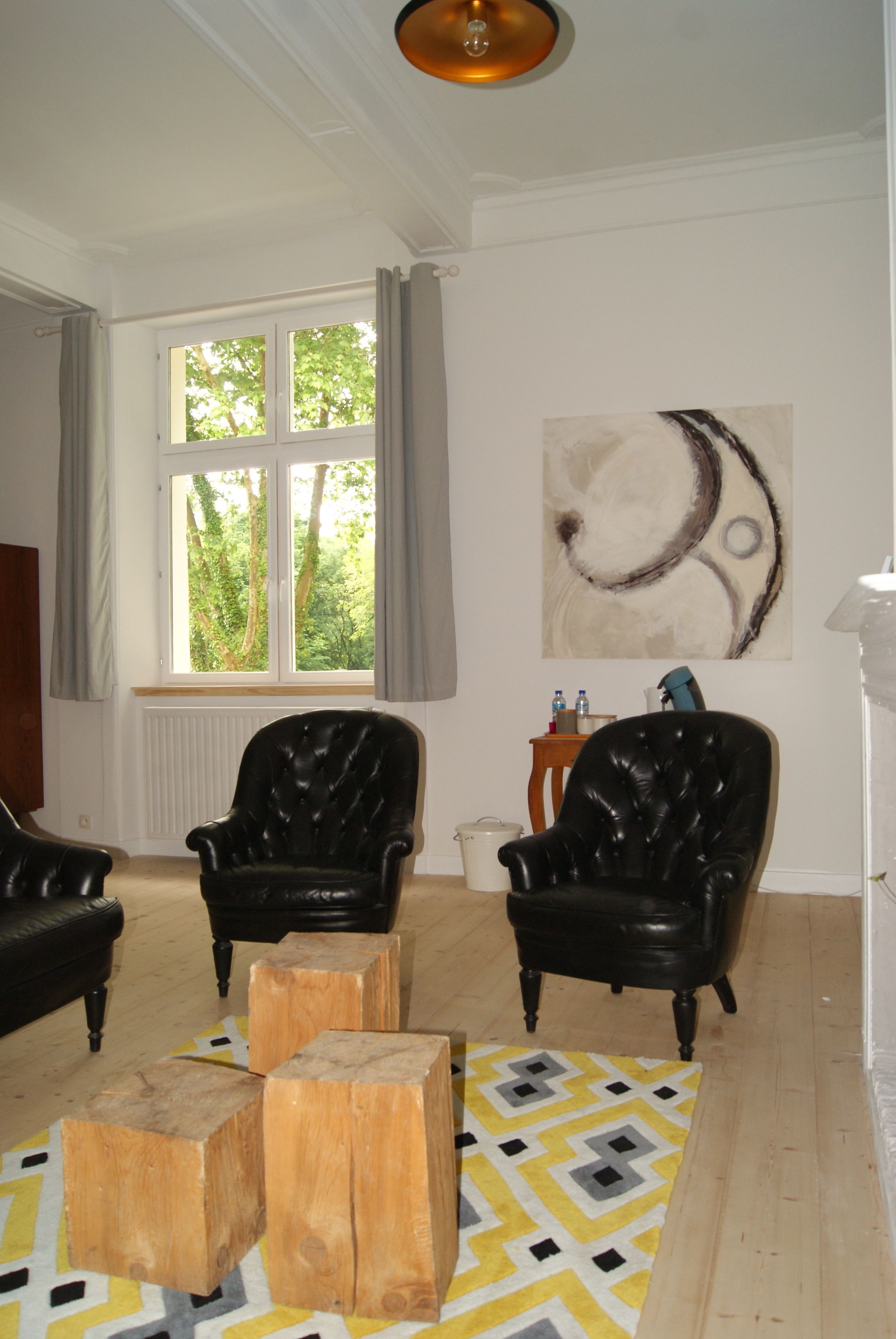 Een woonkamer met twee zwarte leren fauteuils, een houten bijzettafeltje en een geel tapijt met een patroon. Er is een raam met gordijnen, een radiator en een schilderij aan de muur.