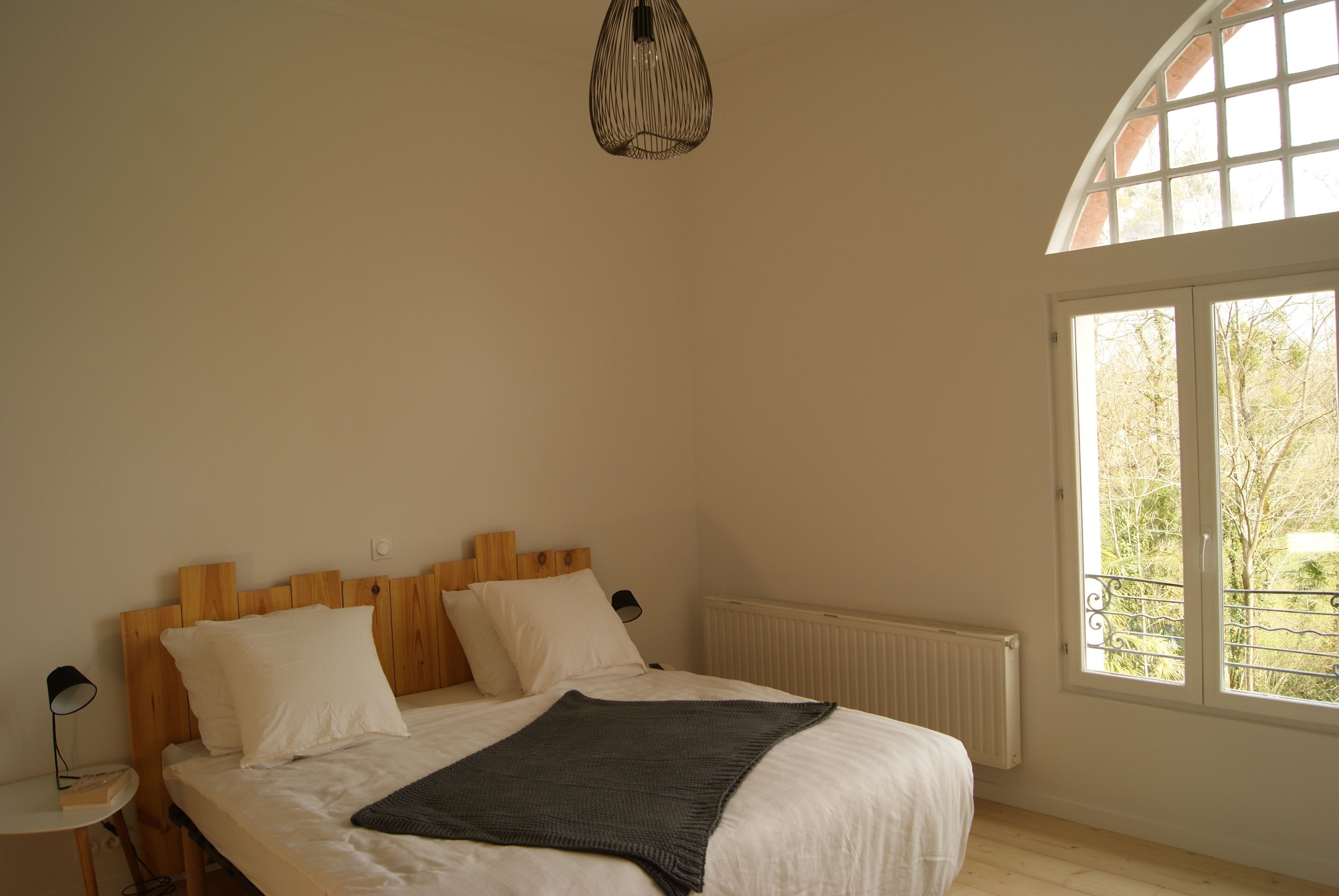 Une chambre à coucher avec une tête de lit en bois, un lit recouvert de linge de lit blanc, une petite table de chevet avec une lampe et une fenêtre donnant sur des arbres.