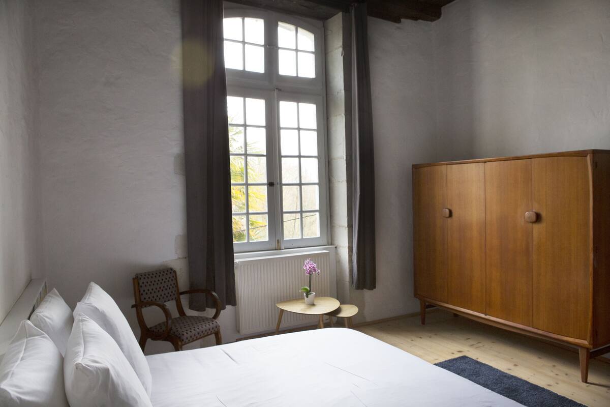 A bedroom with a bed, a wooden wardrobe, a chair, a small table with a flower, and a window with curtains.