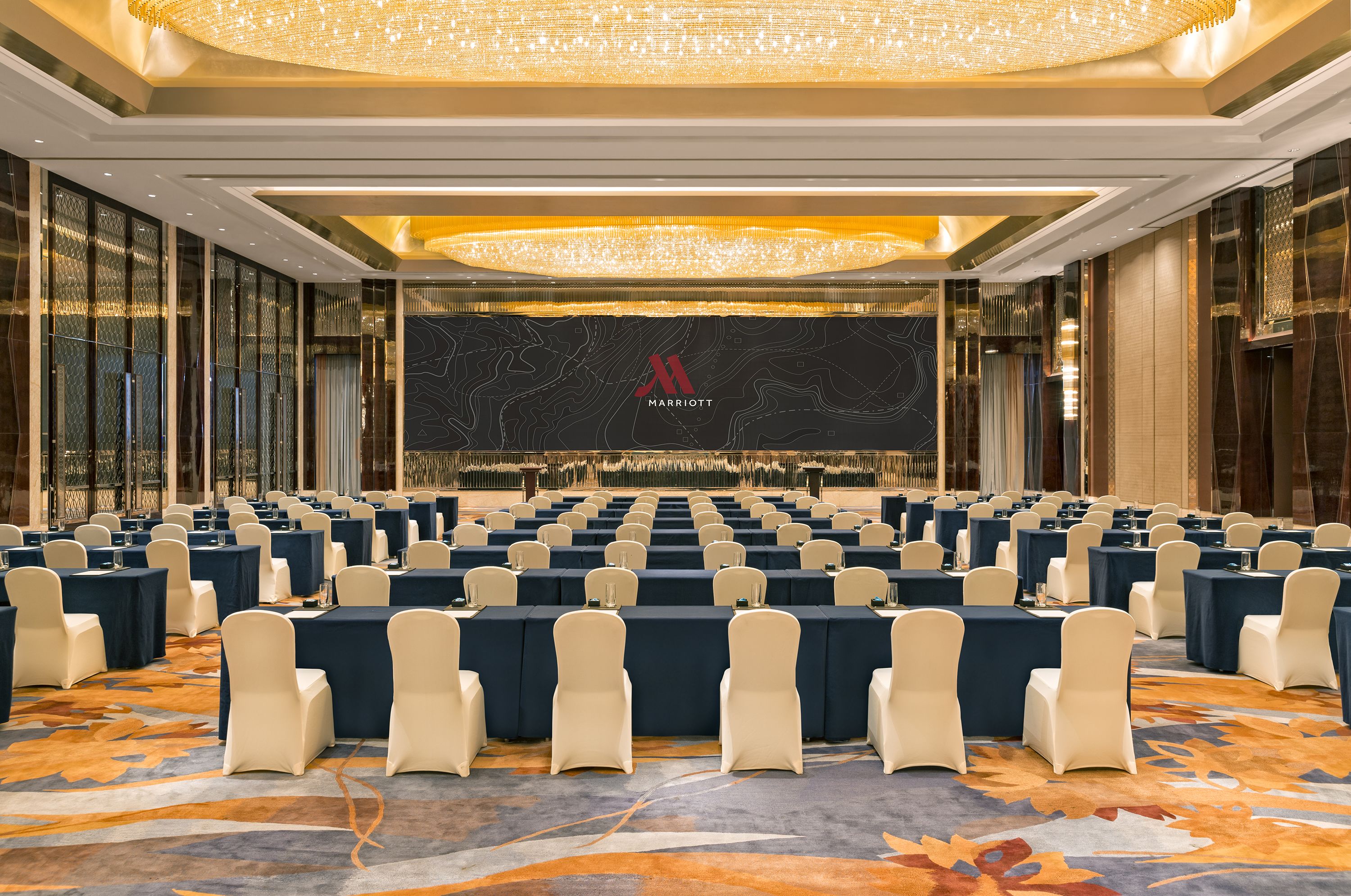 A conference room with rows of tables and chairs, a large chandelier, and a 'Marriott' logo on the wall.