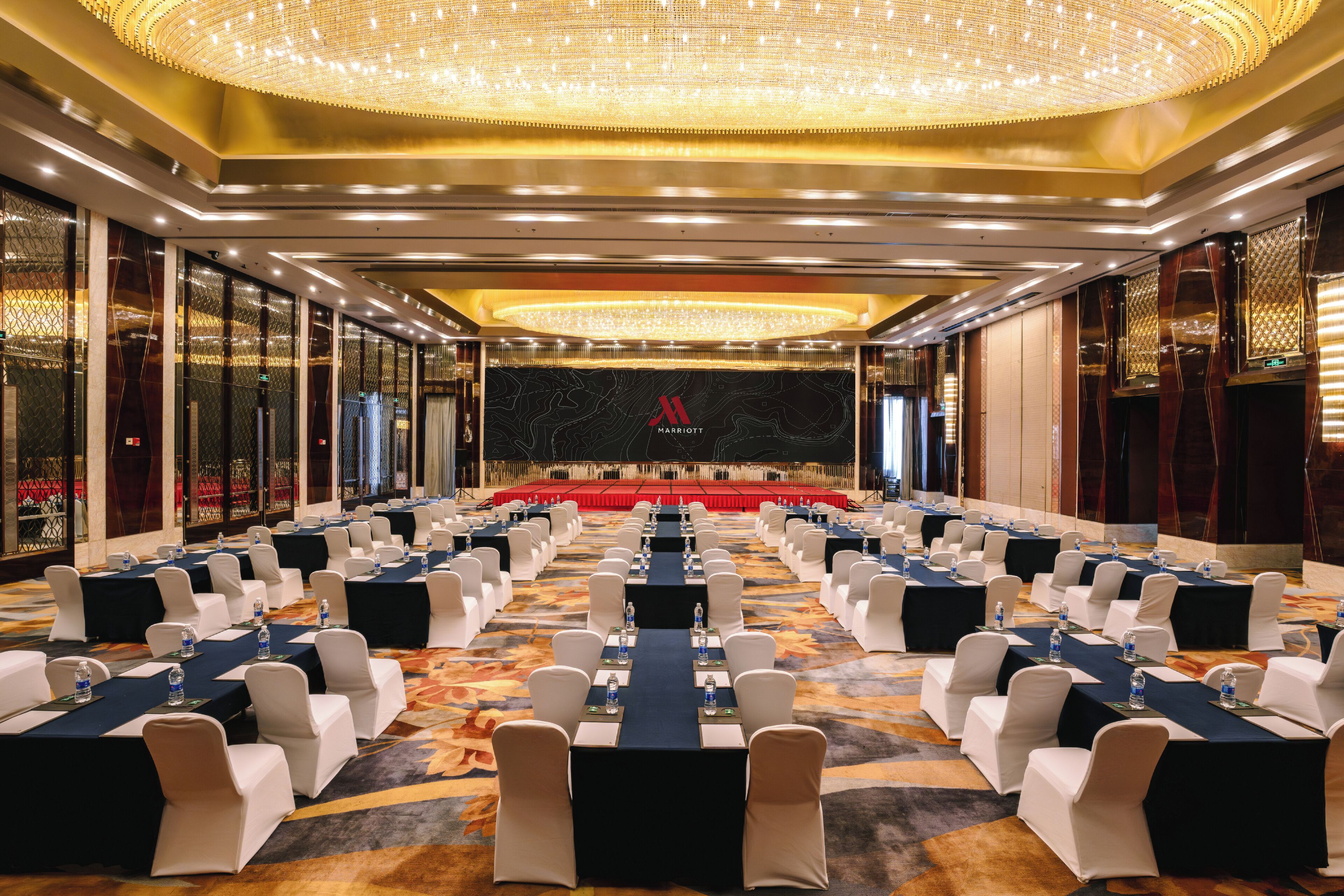 A conference room with rows of tables and chairs, a stage with a red backdrop, and a chandelier.