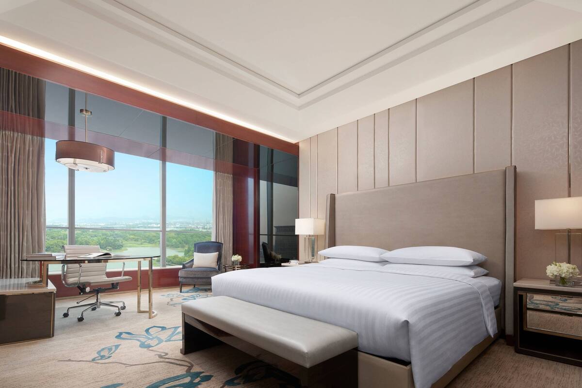 A hotel room with a large bed, a desk, a chair, a bench, and a view of the outdoors.