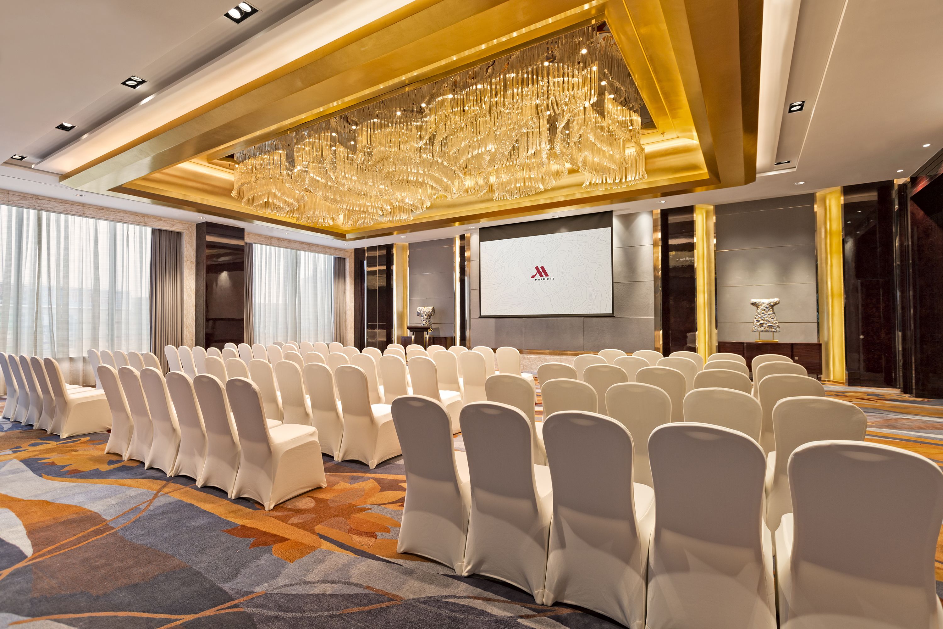 Une salle de conférence avec des rangées de chaises blanches, un grand lustre et un écran de projection.