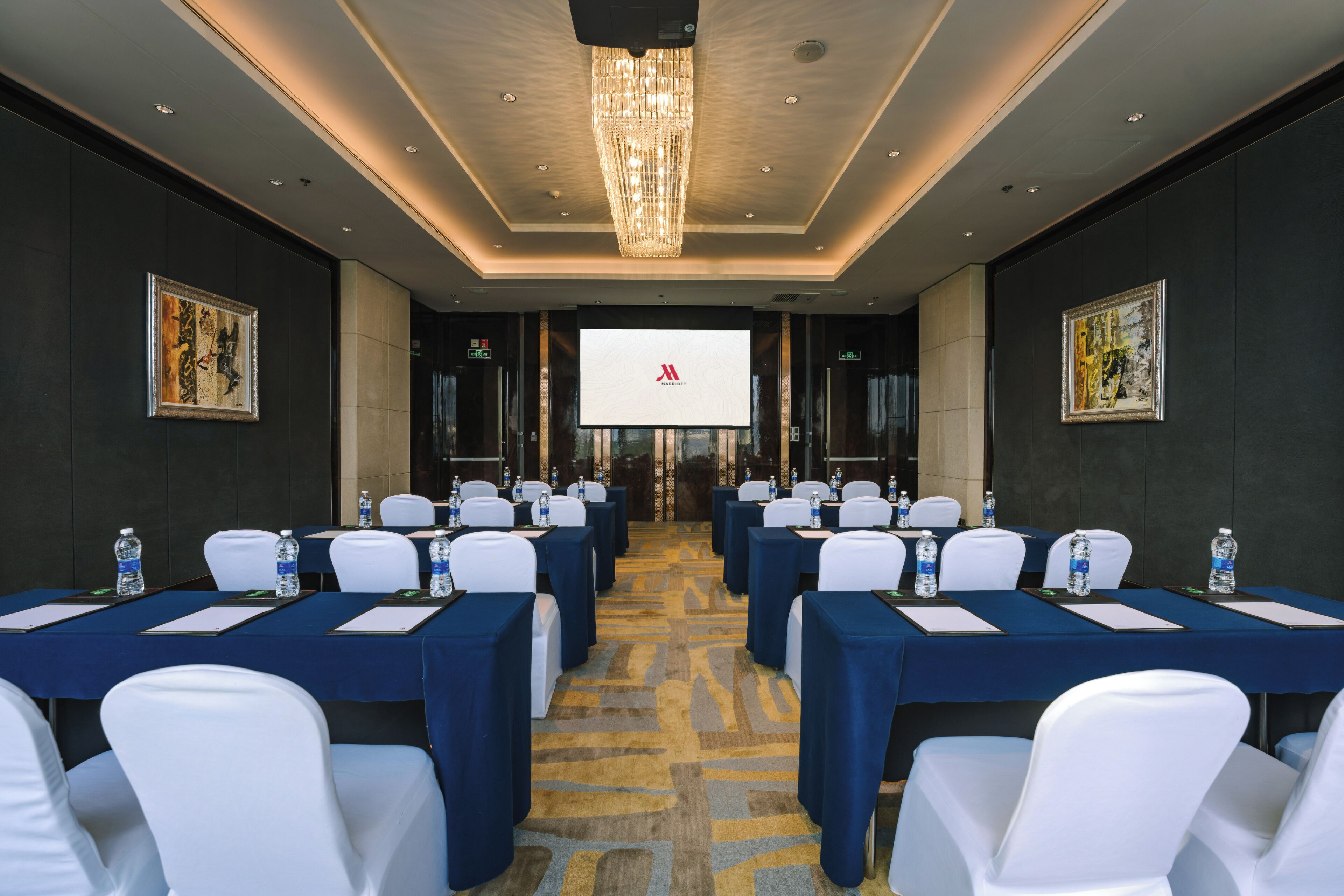 A conference room with a large screen at the front, multiple tables with chairs, and water bottles on each table.