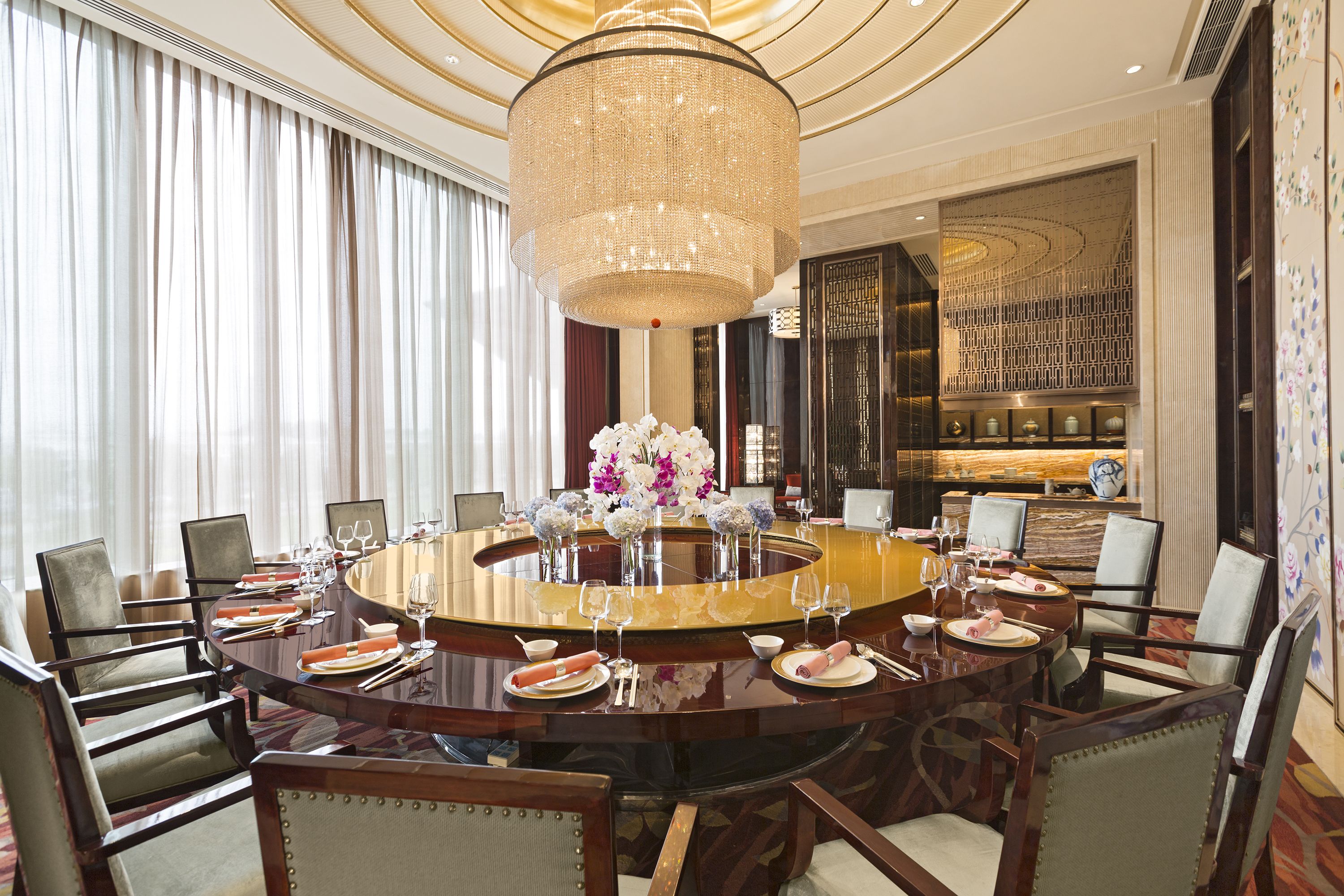 Une table de salle à manger ronde, entourée de chaises, surmontée d’un lustre, et un mur décoratif orné d’un motif.