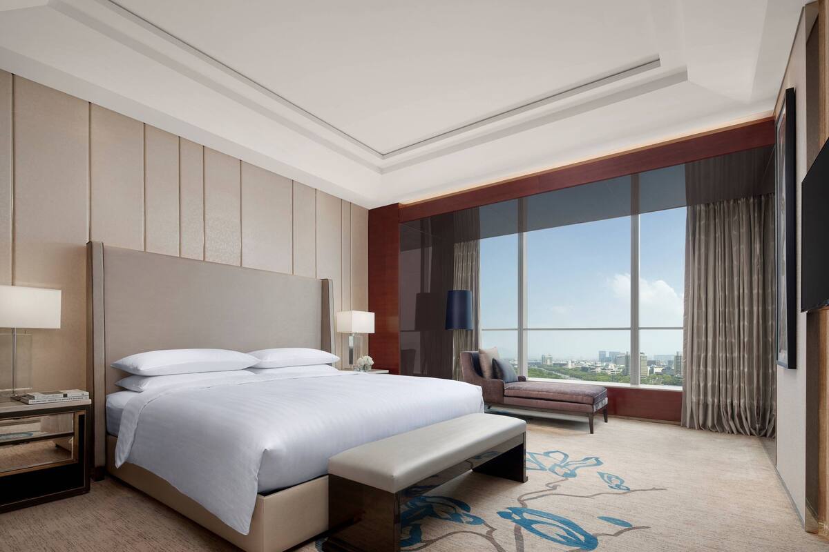 A modern hotel room with a large bed, a bench, a desk with a lamp, and a large window offering a city view.