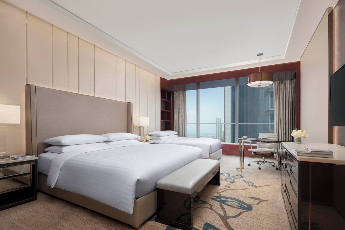 A hotel room with two beds, a desk, a chair, a TV, and a large window with a city view.