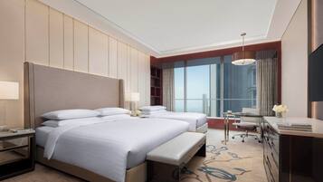 Ein Hotelzimmer mit zwei Betten, einem Schreibtisch, einem Stuhl, einem Fernseher und einem großen Fenster mit Blick auf die Stadt.