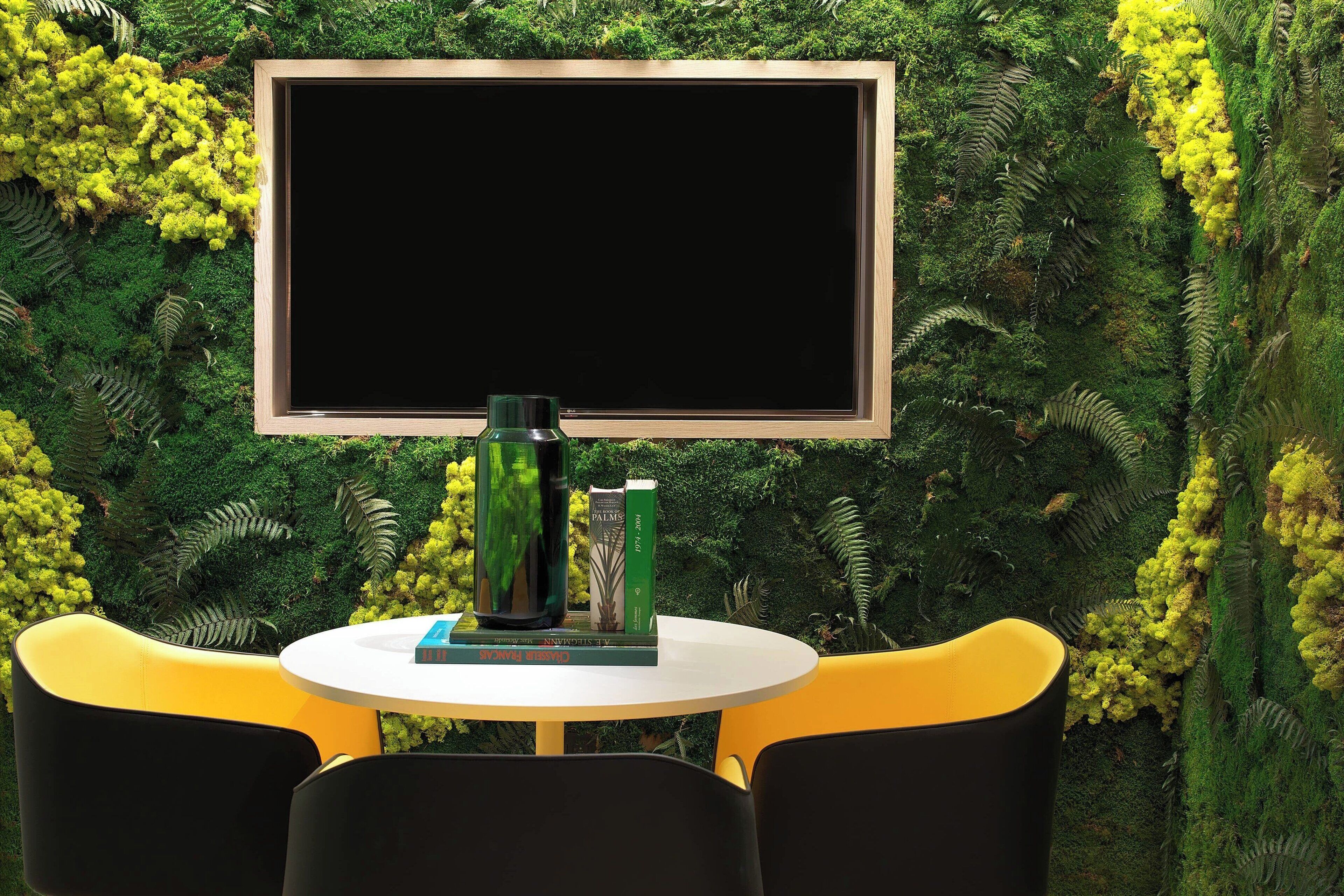 A table with a blank board above it, surrounded by chairs, set against a backdrop of green foliage and yellow flowers.
