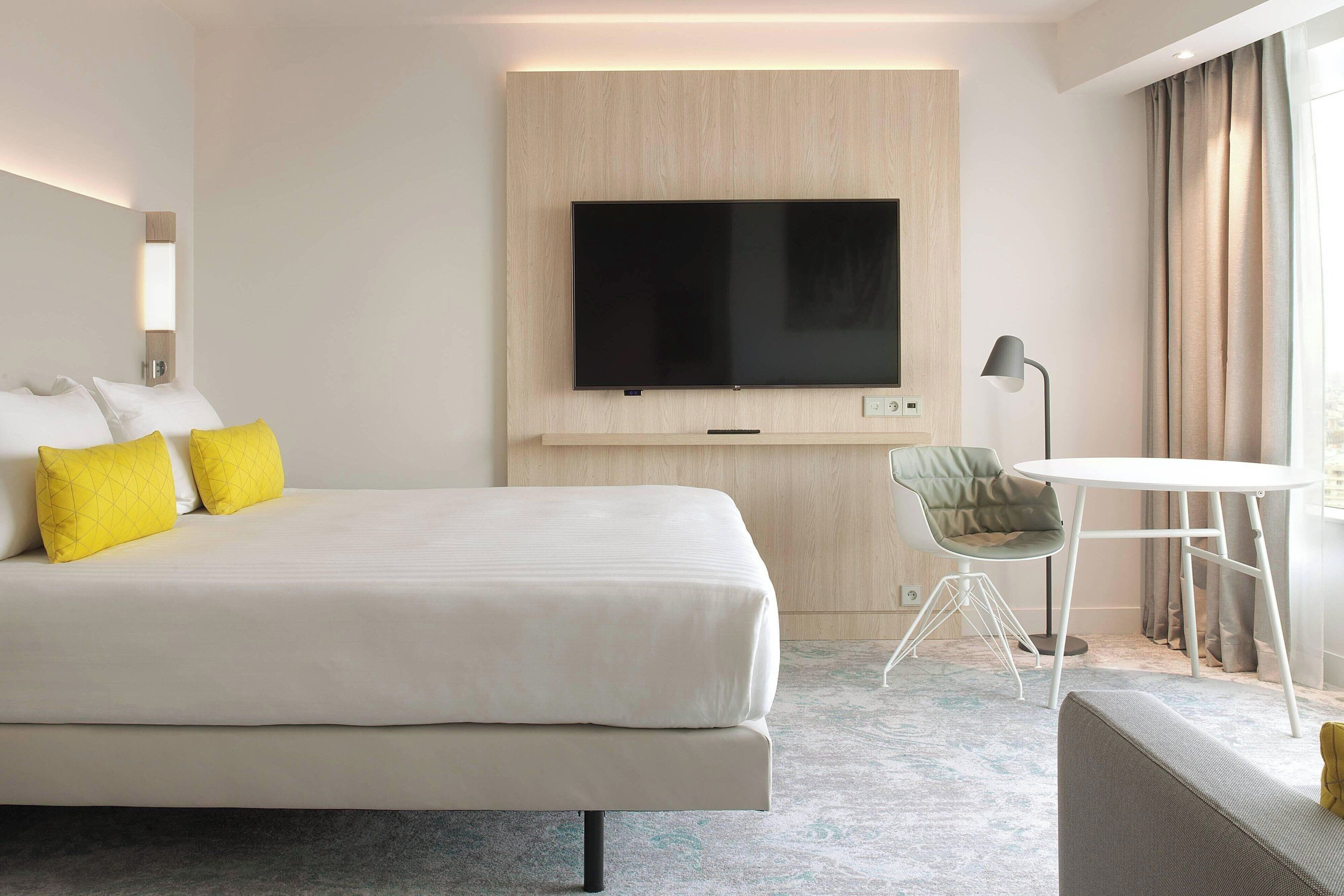 A hotel room with a large bed, a flat-screen TV mounted on the wall, a small round table, a chair, and a lamp.