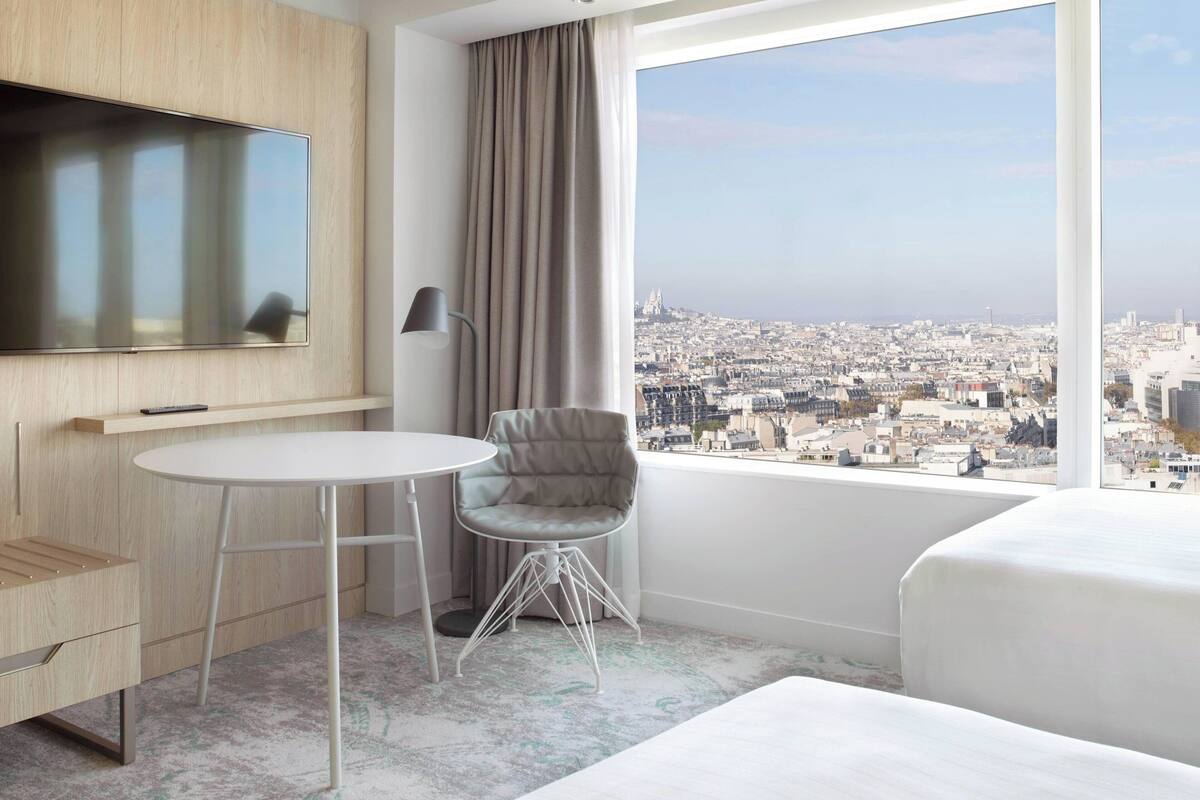 Une chambre d’hôtel avec une grande fenêtre, un lit, un bureau, une chaise et un téléviseur.