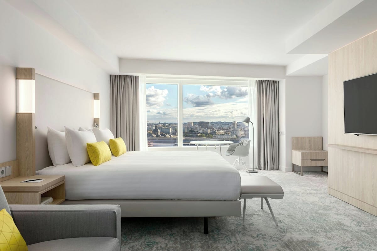 Une chambre d’hôtel moderne dotée d’un grand lit, d’un bureau, d’une chaise, d’une télévision et offrant une vue sur la ville grâce à de grandes fenêtres.