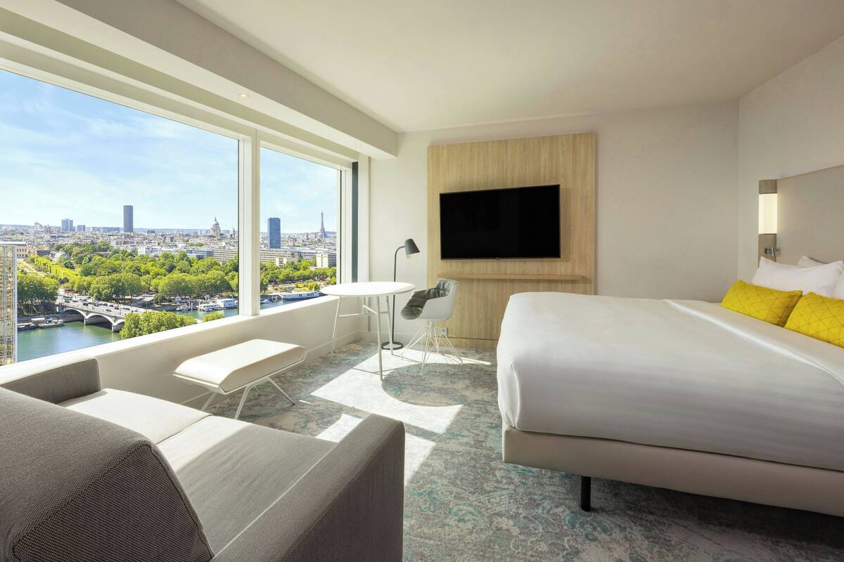 Une chambre d’hôtel avec un grand lit, un canapé, une petite table, un téléviseur fixé au mur et une vue sur le paysage urbain à travers de grandes fenêtres.
