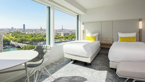 A hotel room with a large window offering a city view, a bed, a bedside table, a chair, and a small table. - Courtyard by  Marriott Paris Gare de Lyon (Paris)