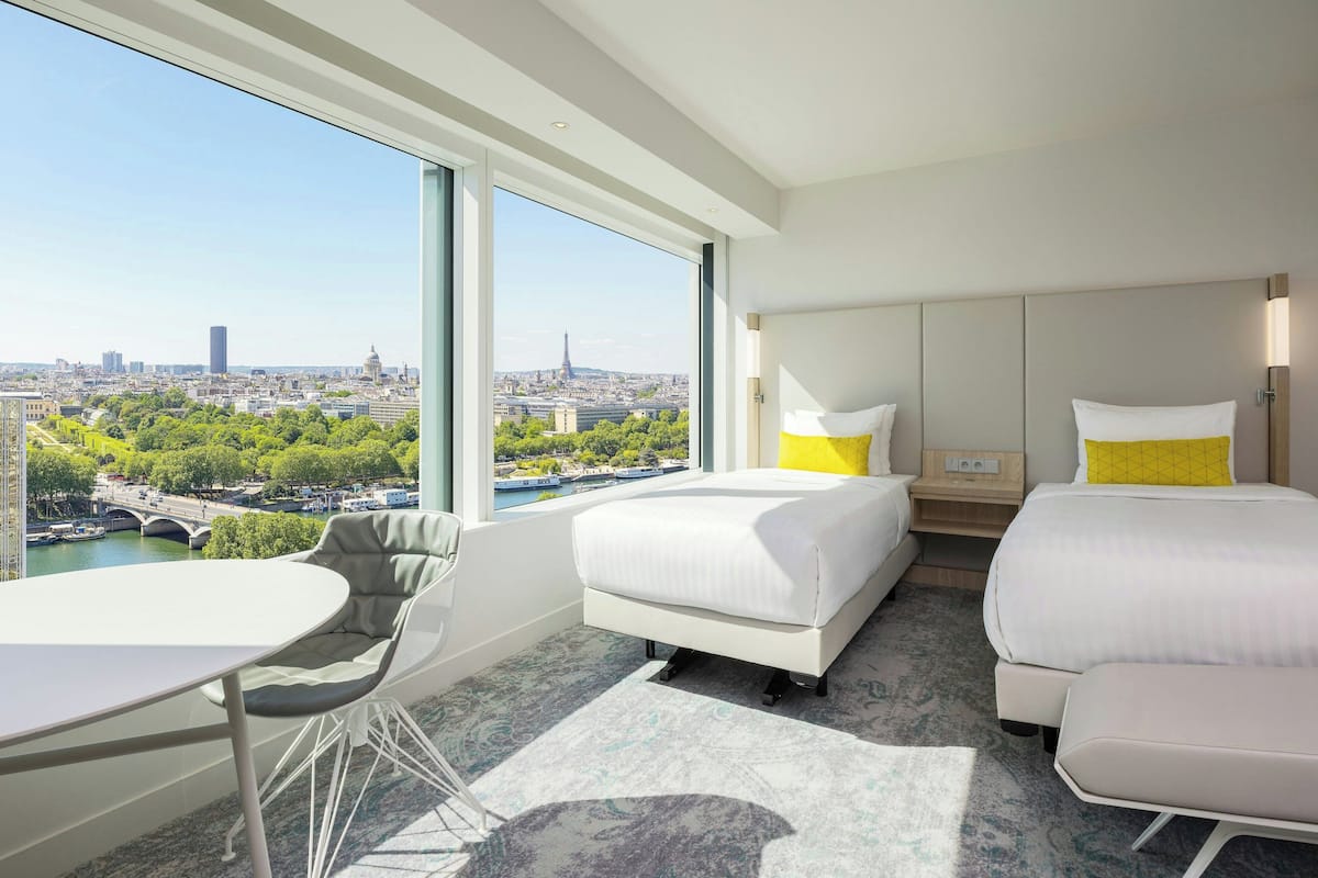 Une chambre d’hôtel dotée d’une grande fenêtre donnant sur la ville, d’un lit, d’une table de chevet, d’une chaise et d’une petite table.