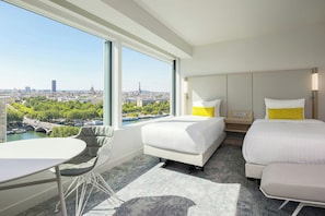 Quarto de hotel com uma janela grande que oferece vista para a cidade, uma cama, uma mesa de cabeceira, uma cadeira e uma pequena mesa.