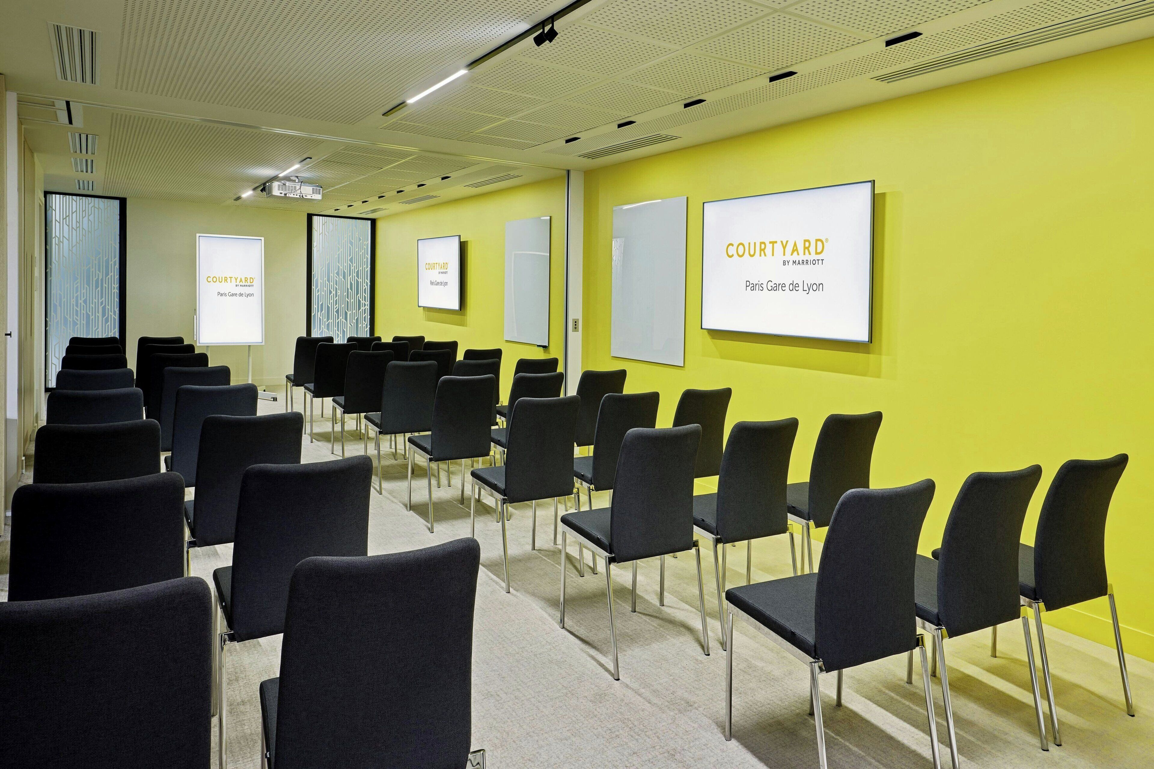 Un salón de conferencias con filas de sillas negras, una pantalla de proyector y un cartel que dice "COURTYARD Paris Gare de Lyon".