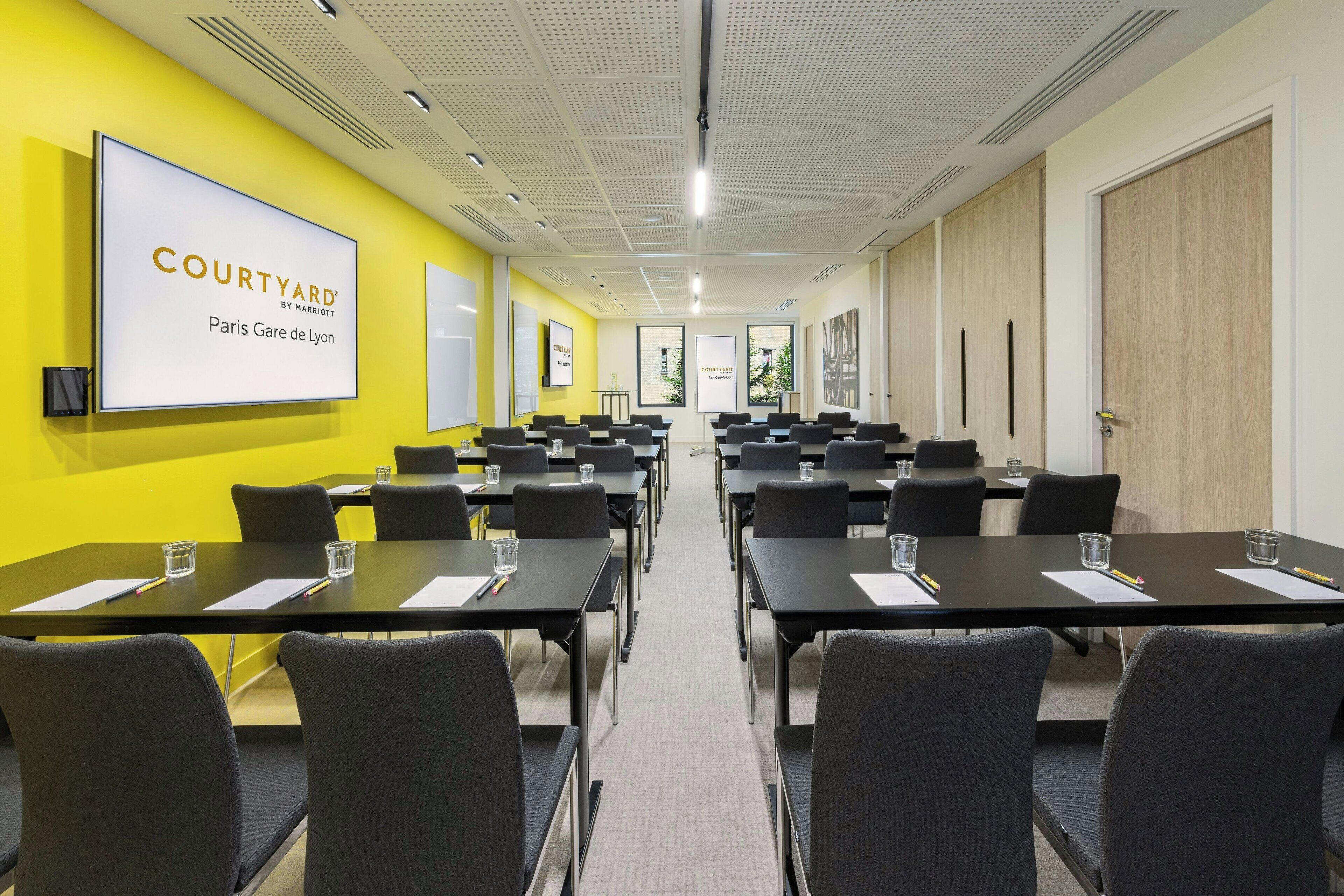 Ein Konferenzraum mit mehreren Tischen und Stühlen, einem großen Bildschirm, der 'COURTYARD PARIS GARE DE LYON' zeigt, und einer leuchtend gelben Wand.