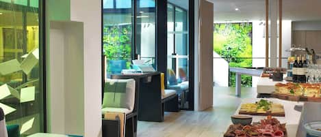 Un espacio interior moderno con una zona de estar, una mesa de buffet con diferentes opciones de comida y vistas a zonas verdes exteriores.