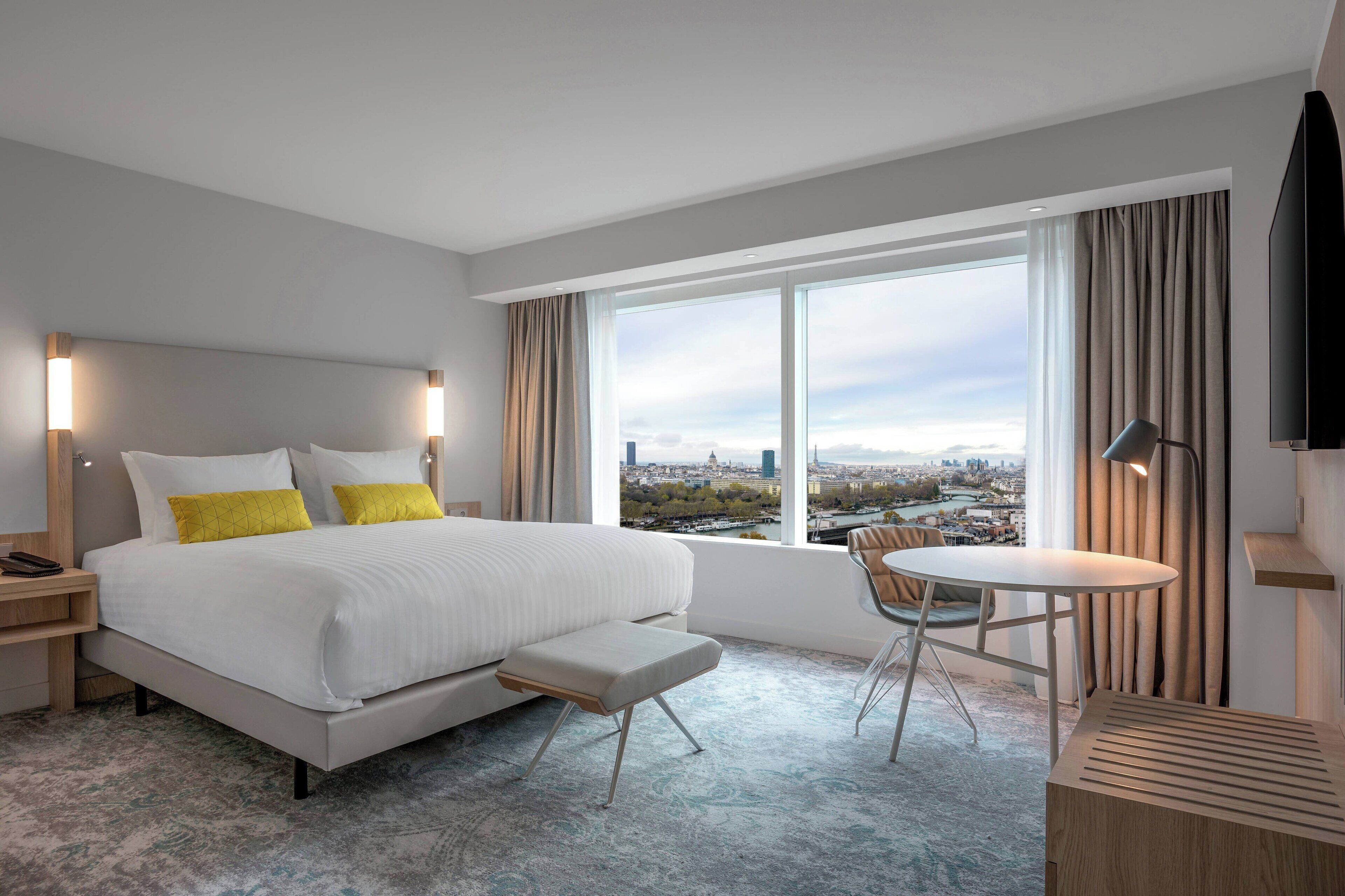 A modern hotel room with a large bed, a small table, and a view of a cityscape.