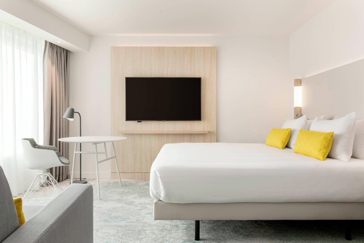 Une chambre d’hôtel moderne avec un grand lit, un téléviseur à écran plat fixé au mur, une petite table ronde et une chaise contemporaine.