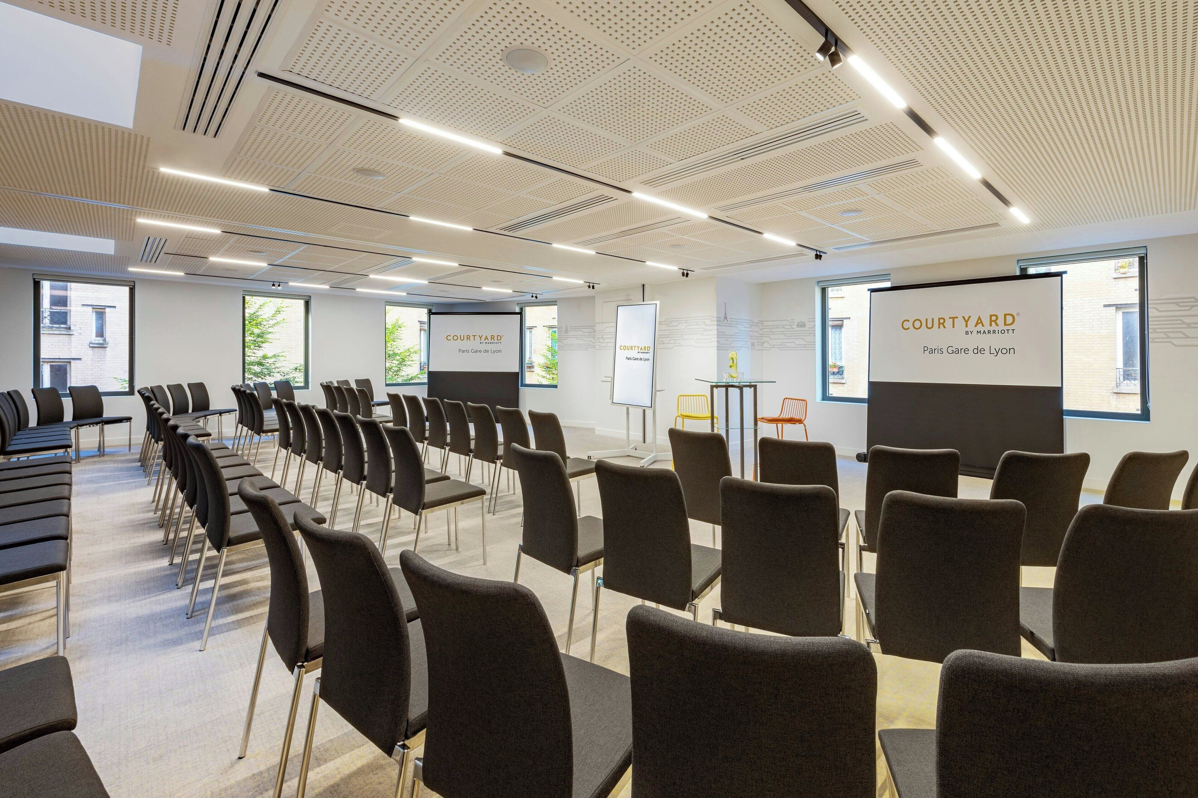 Un salón de conferencias con hileras de sillas, una pantalla de presentación y un cartel que indica la ubicación como Courtyard by Marriott Paris Gare de Lyon.