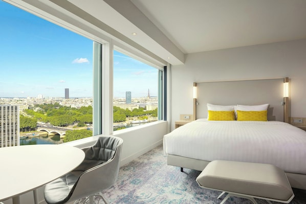 Quarto de hotel com uma janela ampla que oferece vista para a cidade, uma cama com roupa de cama branca e amarela, uma mesinha e uma cadeira.
