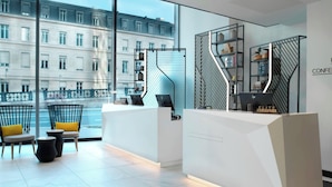 A modern hotel reception area with a white reception desk, seating area with chairs and a small table, and a view of a cityscape through large windows. - Courtyard by  Marriott Paris Gare de Lyon (Paris)