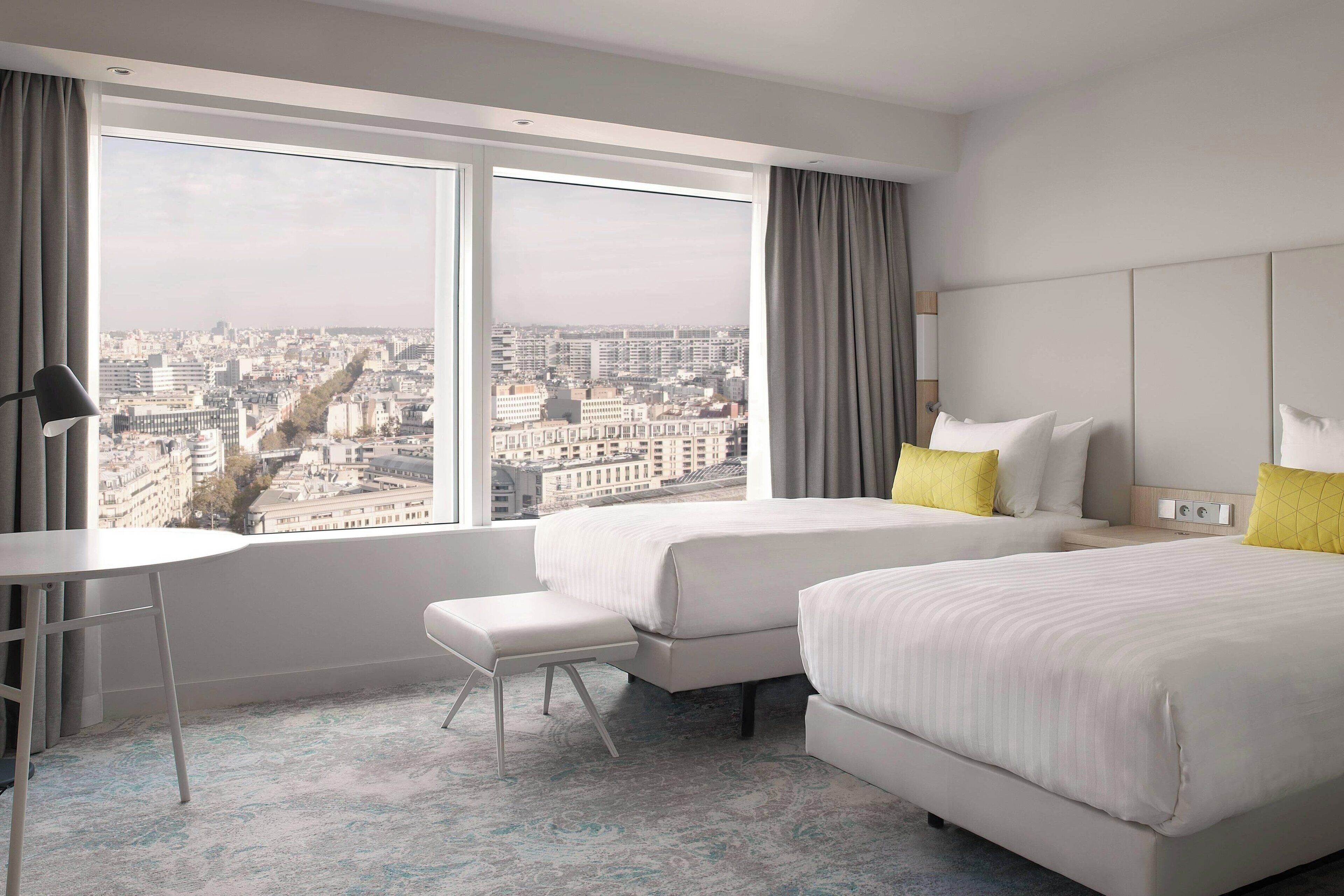 Quarto de hotel com duas camas, uma mesa pequena e um banco. O quarto possui grandes janelas com vista para a cidade.