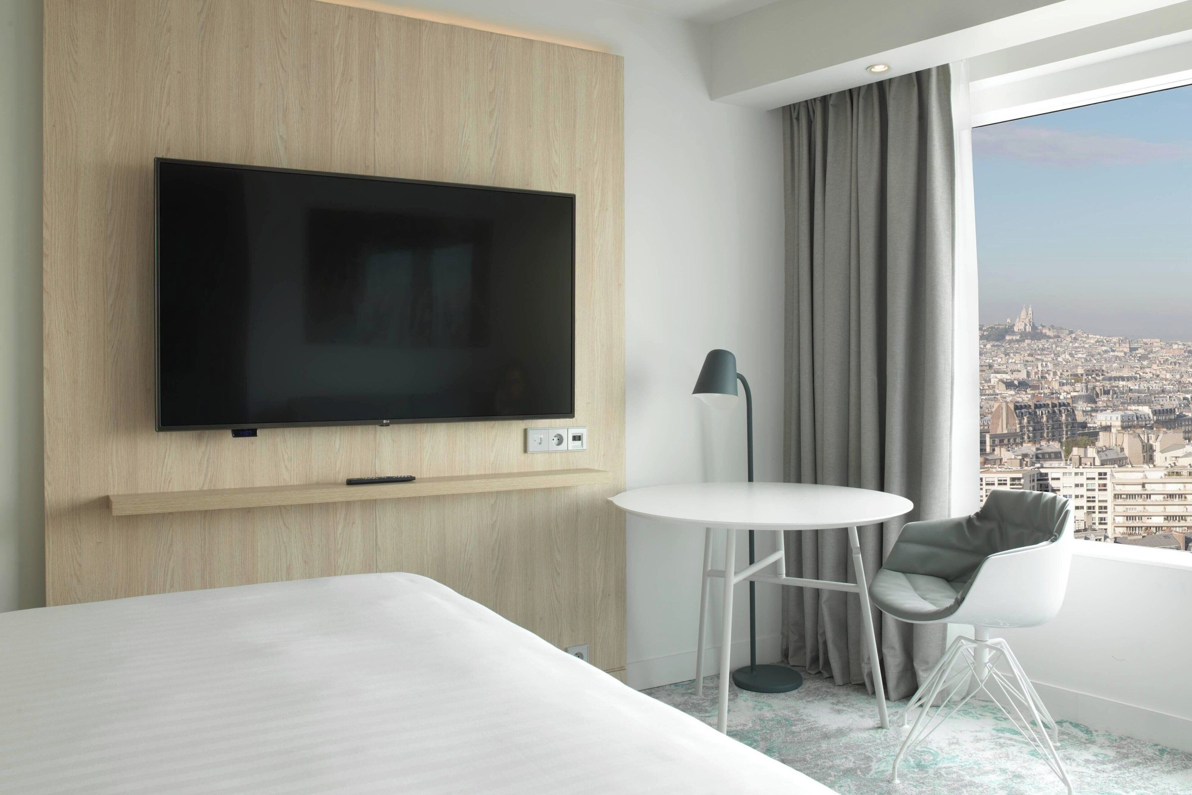 A modern hotel room with a large flat-screen TV mounted on the wall, a round white table, a grey chair, and a view of a cityscape through a large window.