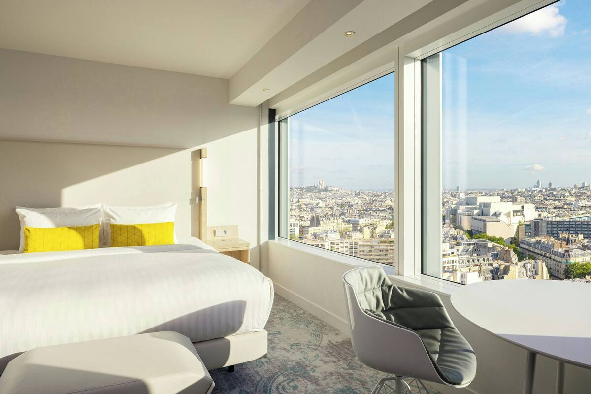 Une chambre d’hôtel moderne dotée d’un grand lit, d’un petit bureau et d’une chaise, et offrant une vue sur la ville grâce à de grandes fenêtres.