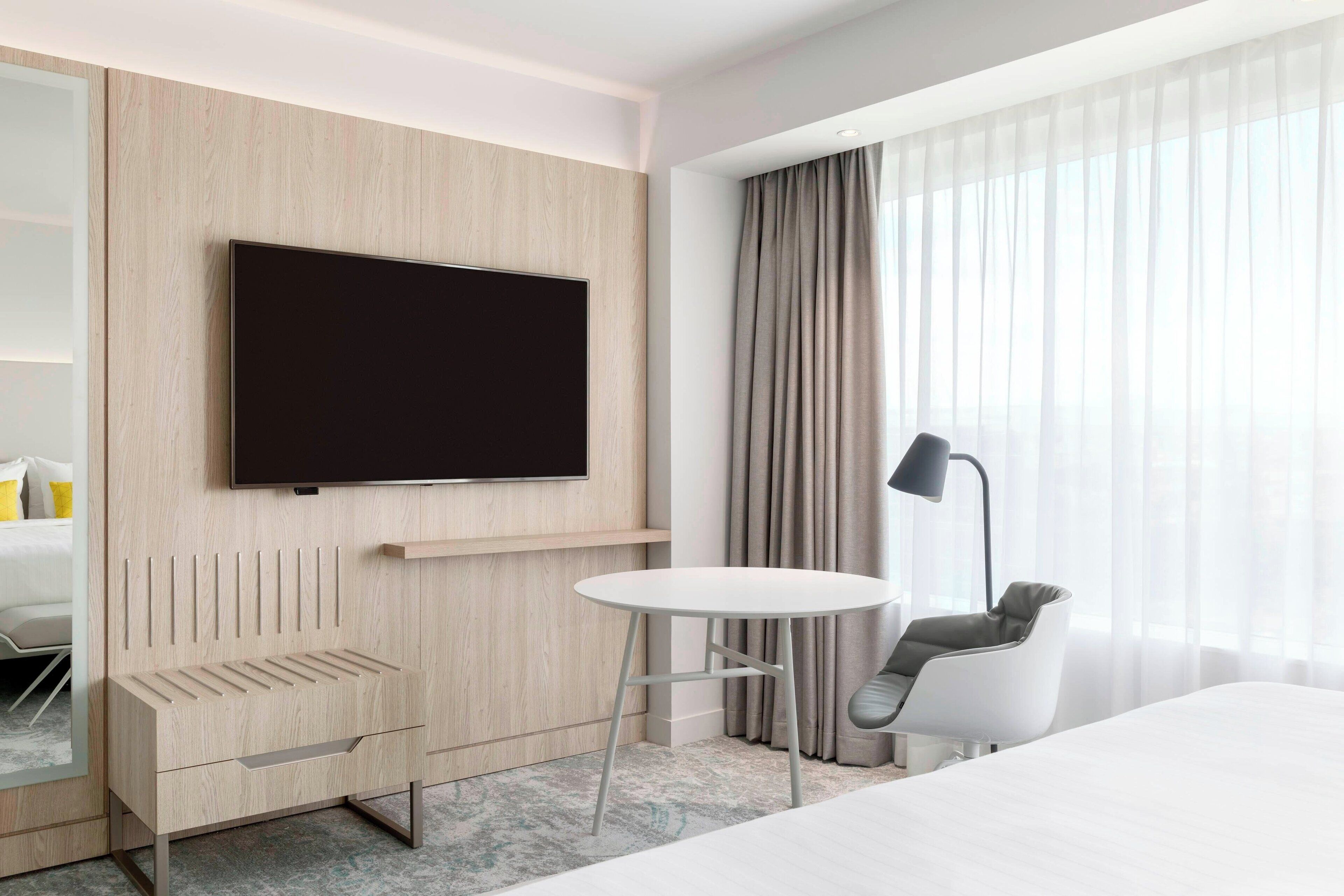A modern hotel room with a large flat-screen TV mounted on a wooden wall, a round white table, a gray chair, and a large window with sheer curtains.
