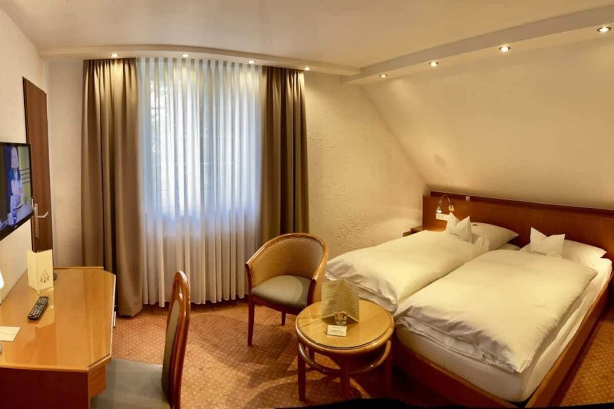 Une chambre d’hôtel comprenant un lit, deux tables de chevet, une chaise, un bureau avec une lampe, une télévision et une fenêtre avec des rideaux.