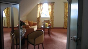 Une chambre d’hôtel comprenant un coin salon, une table à manger et un canapé. On y trouve une fenêtre avec des rideaux, une porte et un miroir.