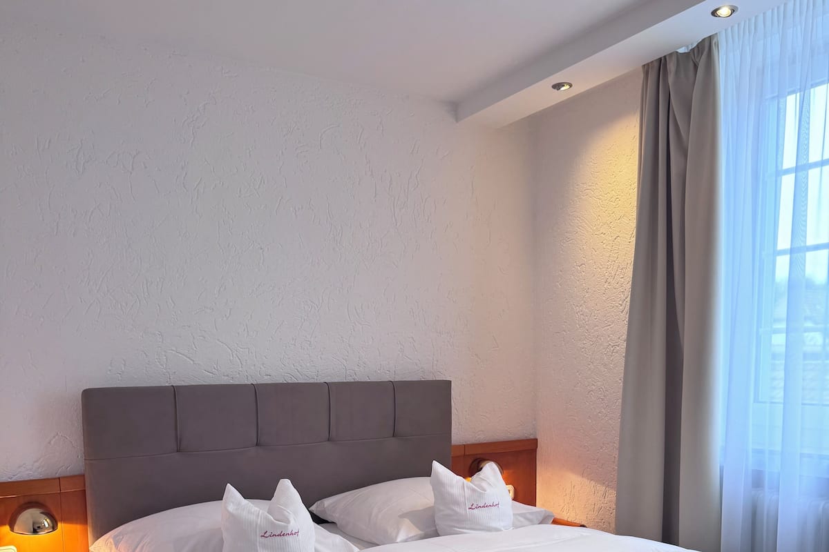 Un lit bien fait, avec du linge de lit blanc et des oreillers, une tête de lit en bois, une table de chevet avec une lampe et une fenêtre avec des rideaux.