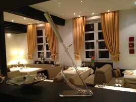 A modern hotel room with a dining area, two sofas, a table with a decorative metal sculpture, and a vase with flowers.